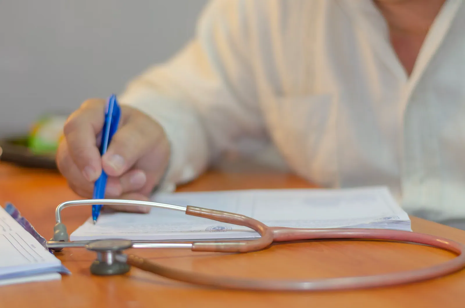 Médico escrevendo um relatório à mesa, com o estetoscópio ao lado
